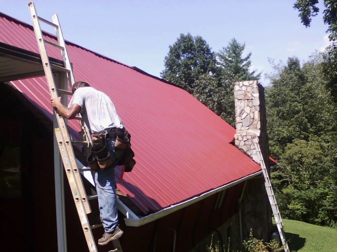 Roofer on a ladder beside a red metal roof and stone chimney — Soffit & Fascia Repair by Roofing Brothers in Warren